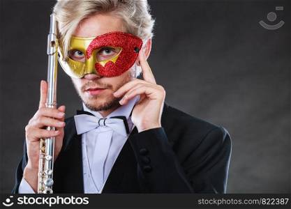 Holidays, people and celebration concept. Elegant young guy wearing suit white shirt bow tie and carnival venetian mask holding flute instrument on dark. man in carnival mask playing flute