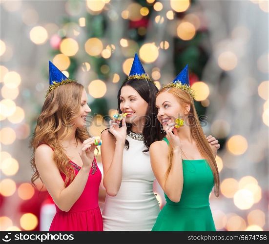 holidays, people and celebration concept - smiling women in party caps blowing to whistles over christmas tree lights background