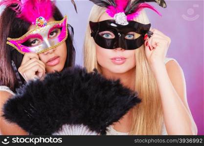 Holidays, people and celebration concept. two women mixed race and caucasian with carnival venetian masks and black feather fan
