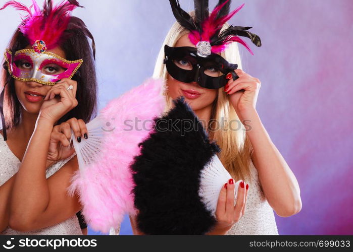 Holidays, people and celebration concept. two women mixed race and caucasian with carnival venetian masks and feather fan