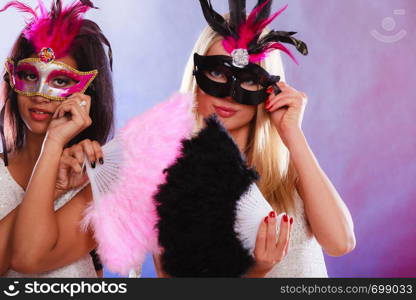Holidays, people and celebration concept. two women mixed race and caucasian with carnival venetian masks and feather fan