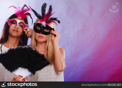 Holidays, people and celebration concept. two women mixed race and caucasian with carnival venetian masks and black feather fan