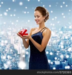 holidays, people and christmas concept - smiling woman in dress holding red gift box over snowy city background