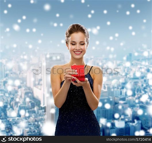holidays, people and christmas concept - smiling woman in dress holding red gift box over snowy city background