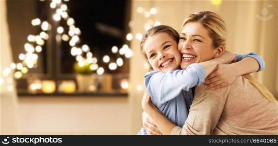 holidays, people and family concept - happy smiling girl with mother hugging on sofa at home over garland lights background. happy smiling family hugging at home on christmas