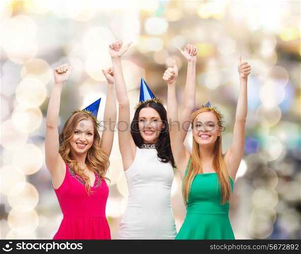 holidays, people, gesture and celebration concept - smiling women in party caps showing thumbs up over lights background