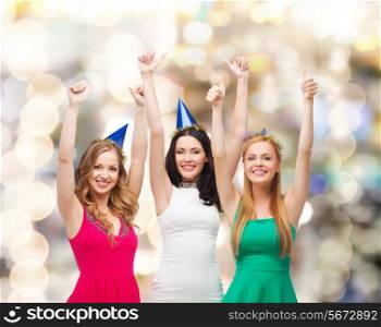 holidays, people, gesture and celebration concept - smiling women in party caps showing thumbs up over lights background