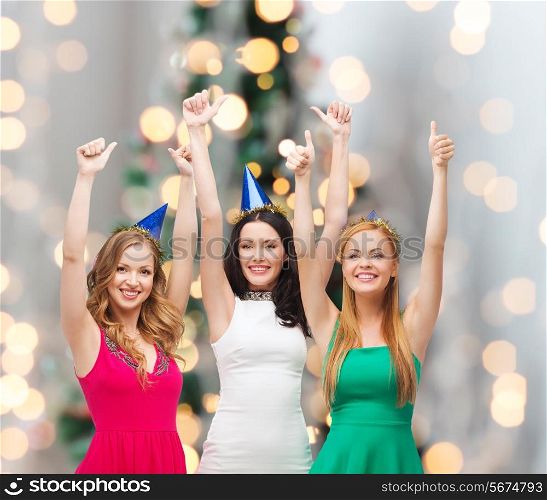 holidays, people, gesture and celebration concept - smiling women in party caps showing thumbs up over christmas tree lights background