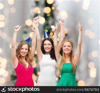 holidays, people, gesture and celebration concept - smiling women in party caps showing thumbs up over christmas tree lights background