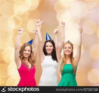 holidays, people, gesture and celebration concept - smiling women in party caps showing thumbs up over beige lights background