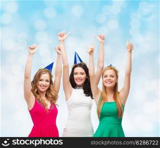 holidays, people, gesture and celebration concept - smiling women in party caps showing thumbs up over blue lights background