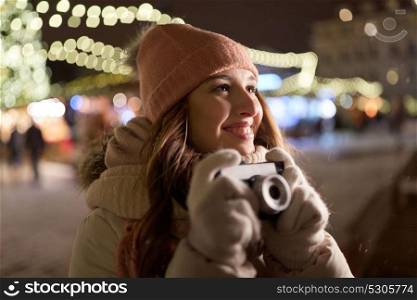 holidays, photography and people concept - beautiful happy young woman with camera at christmas market in winter evening. happy young woman with camera at christmas market