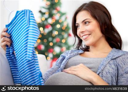 holidays, pregnancy, people and kids clothing concept - happy woman holding and looking at blue baby boys bodysuit over christmas tree background