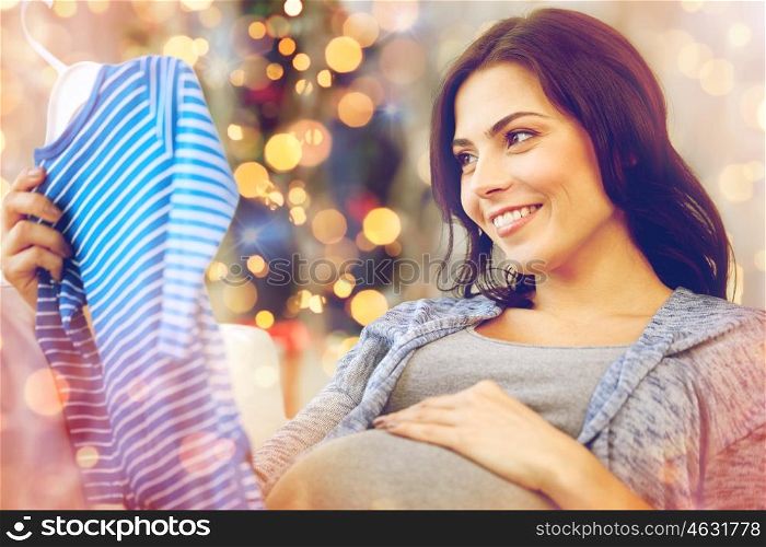 holidays, pregnancy, people and kids clothing concept - happy woman holding and looking at blue baby boys bodysuit over christmas lights background
