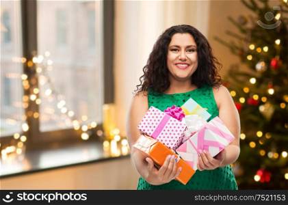 holidays, presents and people concept - happy woman in green dress holding gift boxes over christmas tree lights on home background. happy woman holding gifts over christmas tree