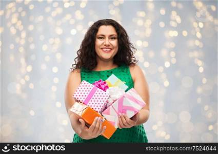 holidays, presents and people concept - happy woman in green dress holding gift boxes over festive lights on grey background. happy woman holding gift boxes over lights