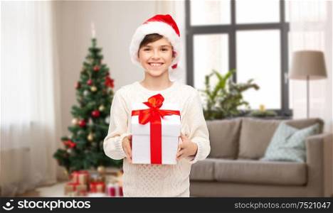 holidays, presents and people concept - smiling happy boy in santa hat with gift box over christmas tree on home background. happy boy in santa hat with christmas gift at home
