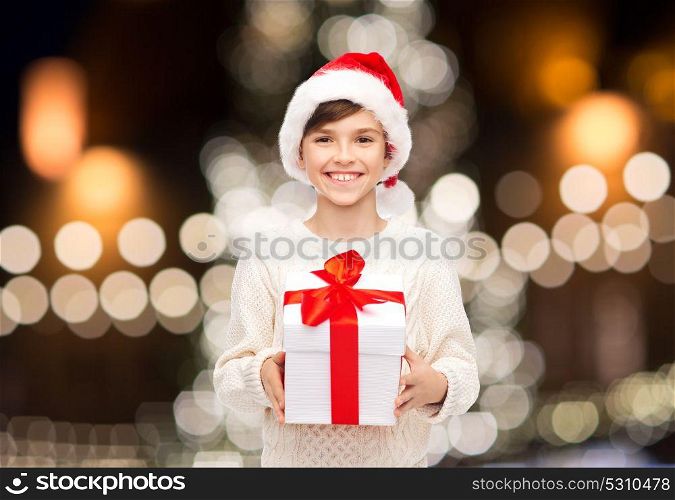 holidays, presents, childhood and people concept - smiling happy boy in santa hat with gift box over christmas tree lights background. smiling happy boy in santa hat with christmas gift