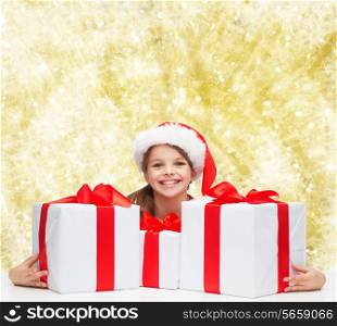 holidays, presents, christmas, childhood and people concept - smiling girl in santa helper hat with gift boxes over yellow lights background