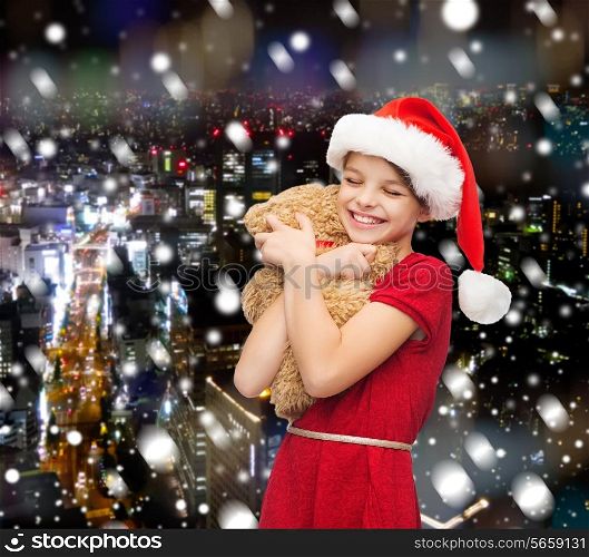 holidays, presents, christmas, childhood and people concept - smiling girl in santa helper hat with teddy bear over snowy night city background