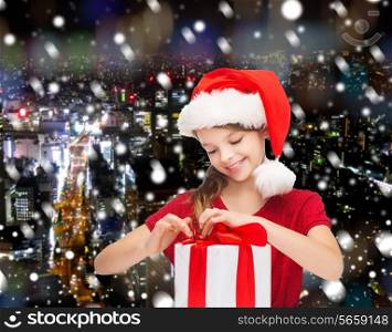 holidays, presents, christmas, childhood and people concept - smiling girl in santa helper hat with gift box over snowing night city background