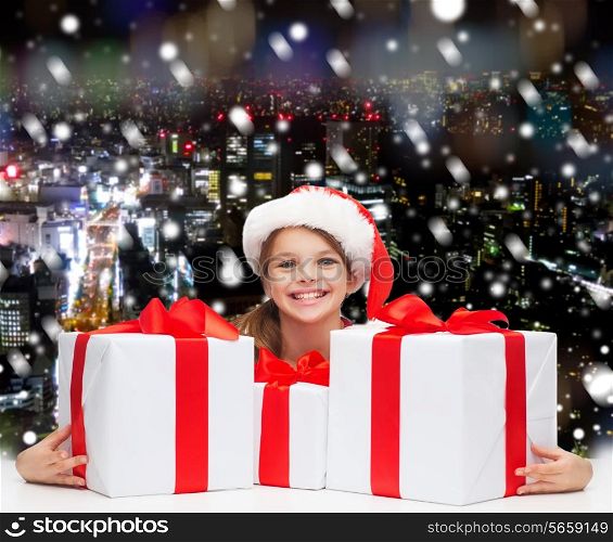holidays, presents, christmas, childhood and people concept - smiling girl in santa helper hat with gift boxes over snowing night city background