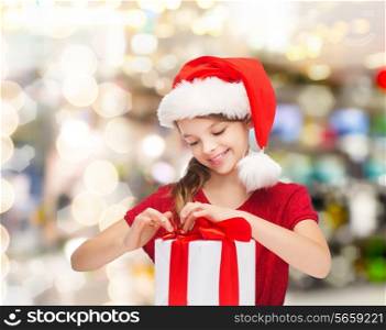 holidays, presents, christmas, childhood and people concept - smiling girl in santa helper hat with gift box over lights background