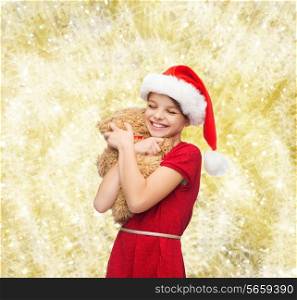 holidays, presents, christmas, childhood and people concept - smiling girl in santa helper hat with teddy bear over yellow lights background