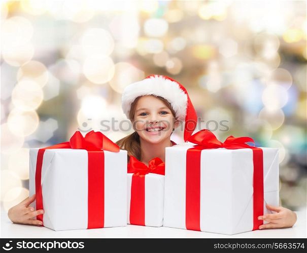 holidays, presents, christmas, childhood and people concept - smiling girl in santa helper hat with gift boxes over lights background