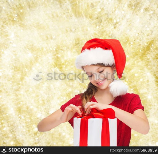 holidays, presents, christmas, childhood and people concept - smiling girl in santa helper hat with gift box over yellow lights background