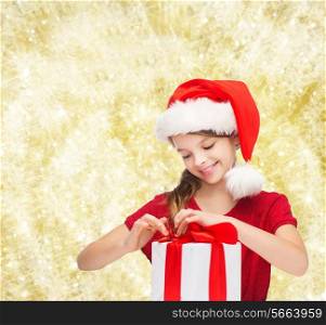 holidays, presents, christmas, childhood and people concept - smiling girl in santa helper hat with gift box over yellow lights background