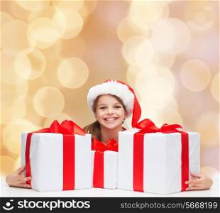 holidays, presents, christmas, childhood and people concept - smiling girl in santa helper hat with gift boxes over beige lights background