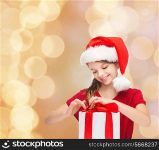 holidays, presents, christmas, childhood and people concept - smiling girl in santa helper hat with gift box over beige lights background