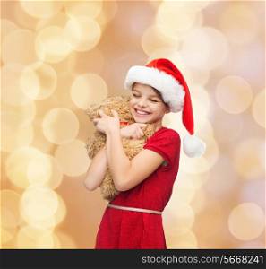 holidays, presents, christmas, childhood and people concept - smiling girl in santa helper hat with teddy bear over beige lights background