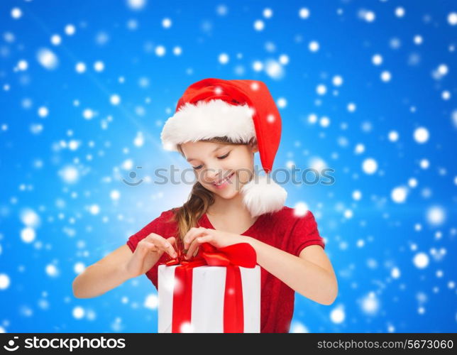 holidays, presents, christmas, childhood and people concept - smiling girl in santa helper hat with gift box over blue snowing background