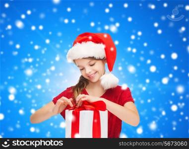 holidays, presents, christmas, childhood and people concept - smiling girl in santa helper hat with gift box over blue snowing background