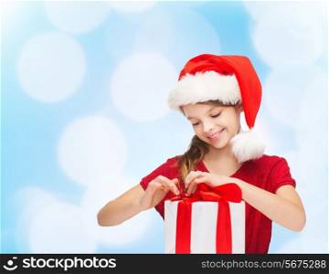 holidays, presents, christmas, childhood and people concept - smiling girl in santa helper hat with gift box over blue lights background