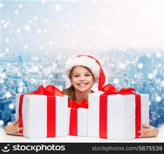 holidays, presents, christmas, childhood and people concept - smiling girl in santa helper hat with gift boxes over snowing city background