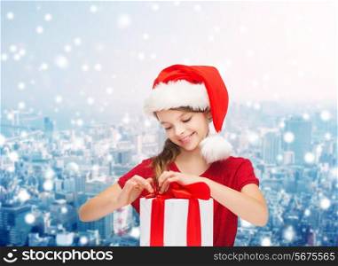 holidays, presents, christmas, childhood and people concept - smiling girl in santa helper hat with gift box over snowing city background