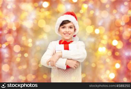 holidays, presents, christmas, childhood and people concept - smiling happy boy in santa hat with gift box over lights background