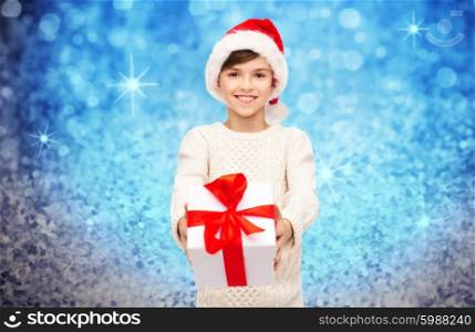 holidays, presents, christmas, childhood and people concept - smiling happy boy in santa hat with gift box over blue lights or glitter background