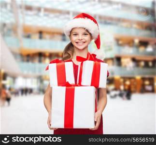holidays, presents, christmas, childhood and people concept - smiling little girl in santa helper hat with gift boxes over shopping center background