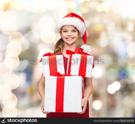 holidays, presents, christmas, childhood and people concept - smiling little girl in santa helper hat with gift boxes over lights background