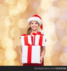 holidays, presents, christmas, childhood and people concept - smiling little girl in santa helper hat with gift boxes over beige lights background