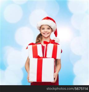 holidays, presents, christmas, childhood and people concept - smiling little girl in santa helper hat with gift boxes over blue lights background