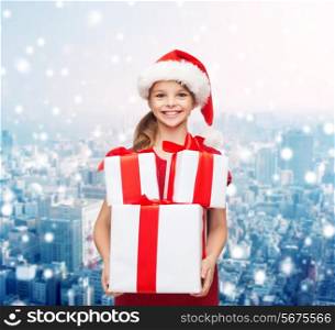 holidays, presents, christmas, childhood and people concept - smiling little girl in santa helper hat with gift boxes over snowy city background
