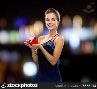holidays, presents, luxury and happiness concept - smiling woman in dress holding red gift box night lights background