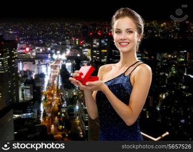 holidays, presents, luxury and happiness concept - smiling woman in dress holding red gift box over night city background