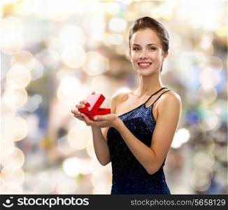 holidays, presents, luxury and happiness concept - smiling woman in dress holding red gift box over lights background