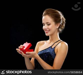 holidays, presents, luxury and happiness concept - smiling woman in dress holding red gift box over black background